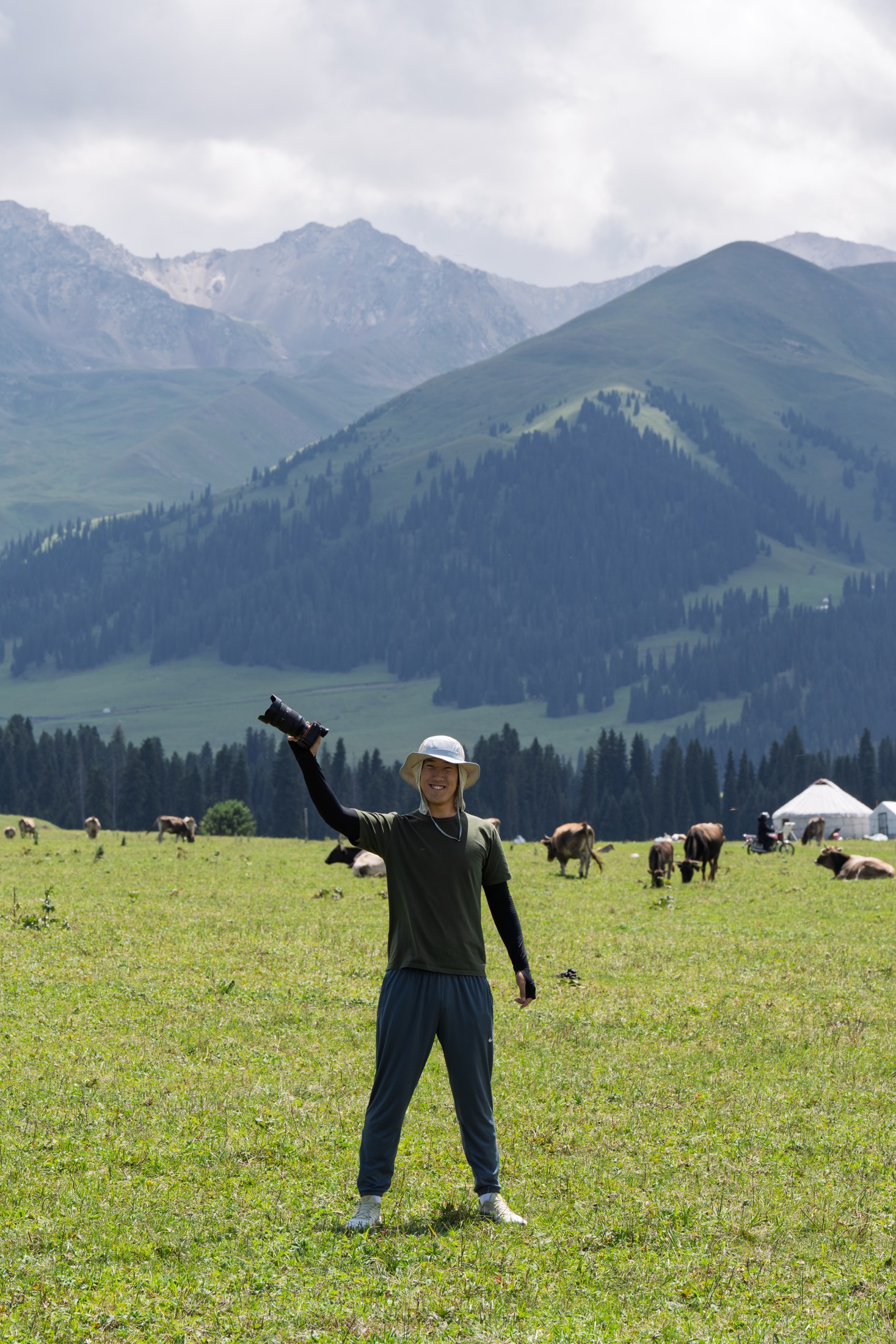 Changzheng Yu smiles and raises his camera in a green mountain meadow, with grazing cattle and forested slopes behind him.