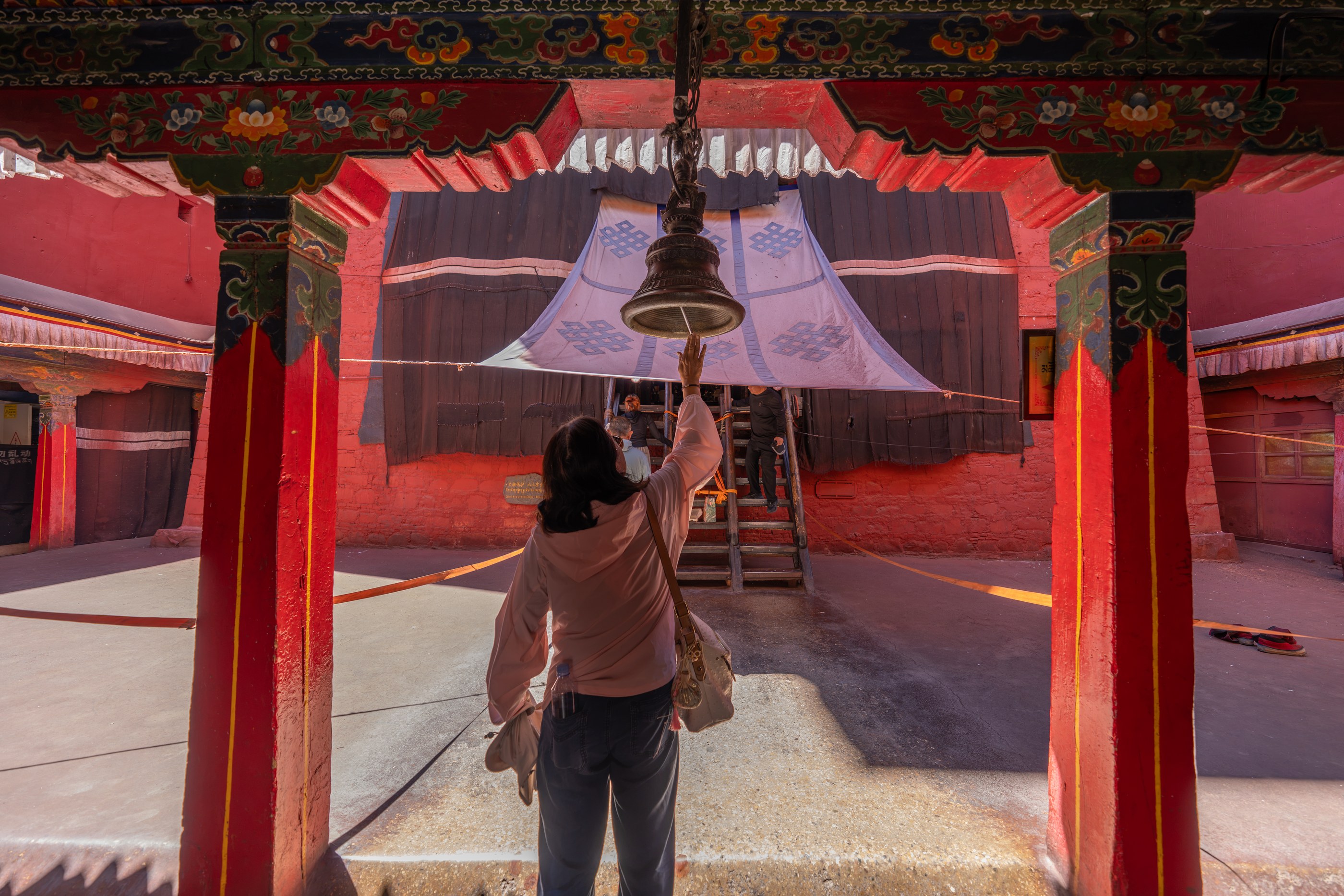 A person reaches toward a hanging bell beneath a decorated red gateway in a sunlit courtyard.
